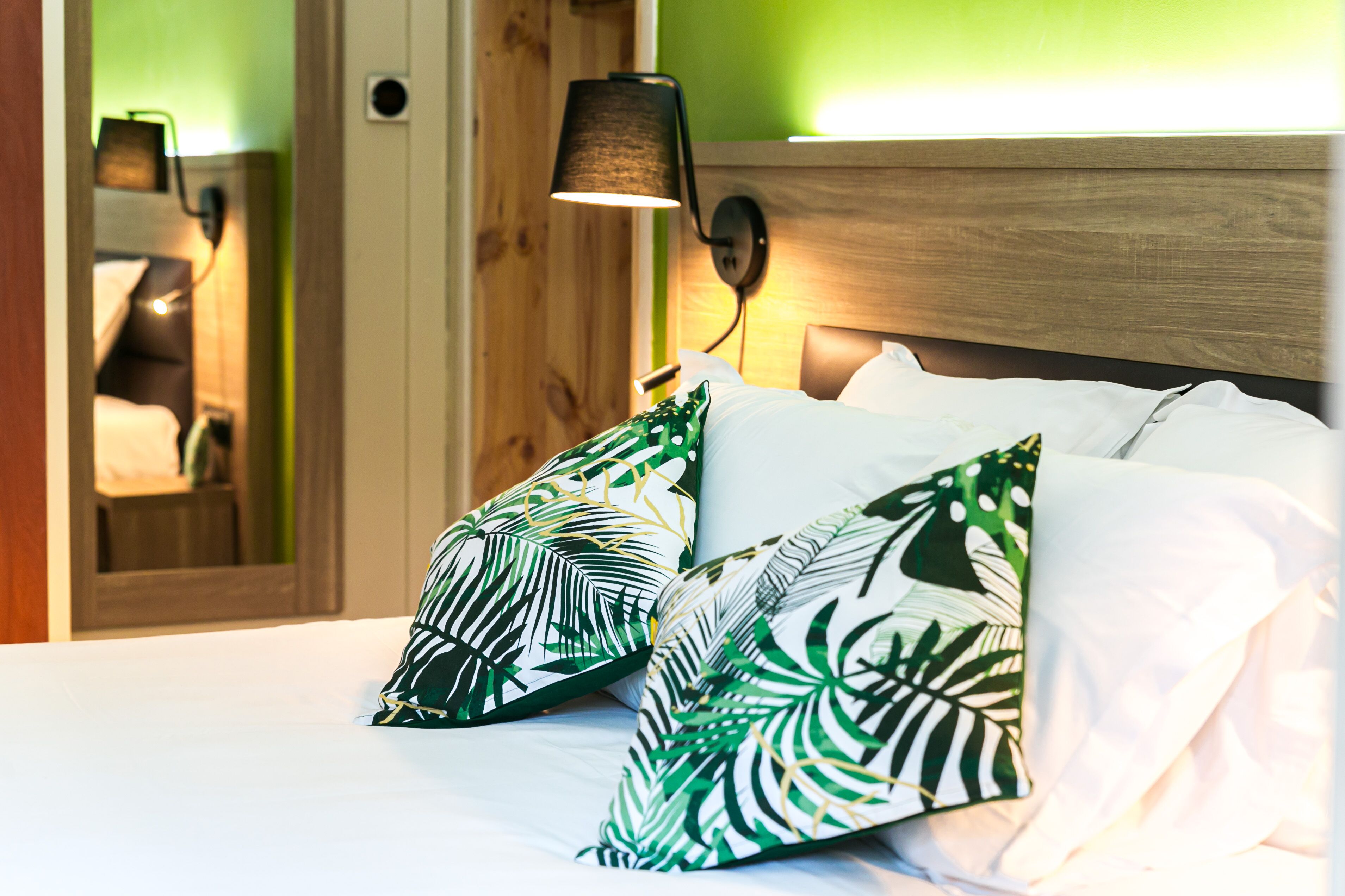 A neatly made bed with white linens and green leaf-patterned pillows, a wooden headboard, and a wall-mounted lamp.
