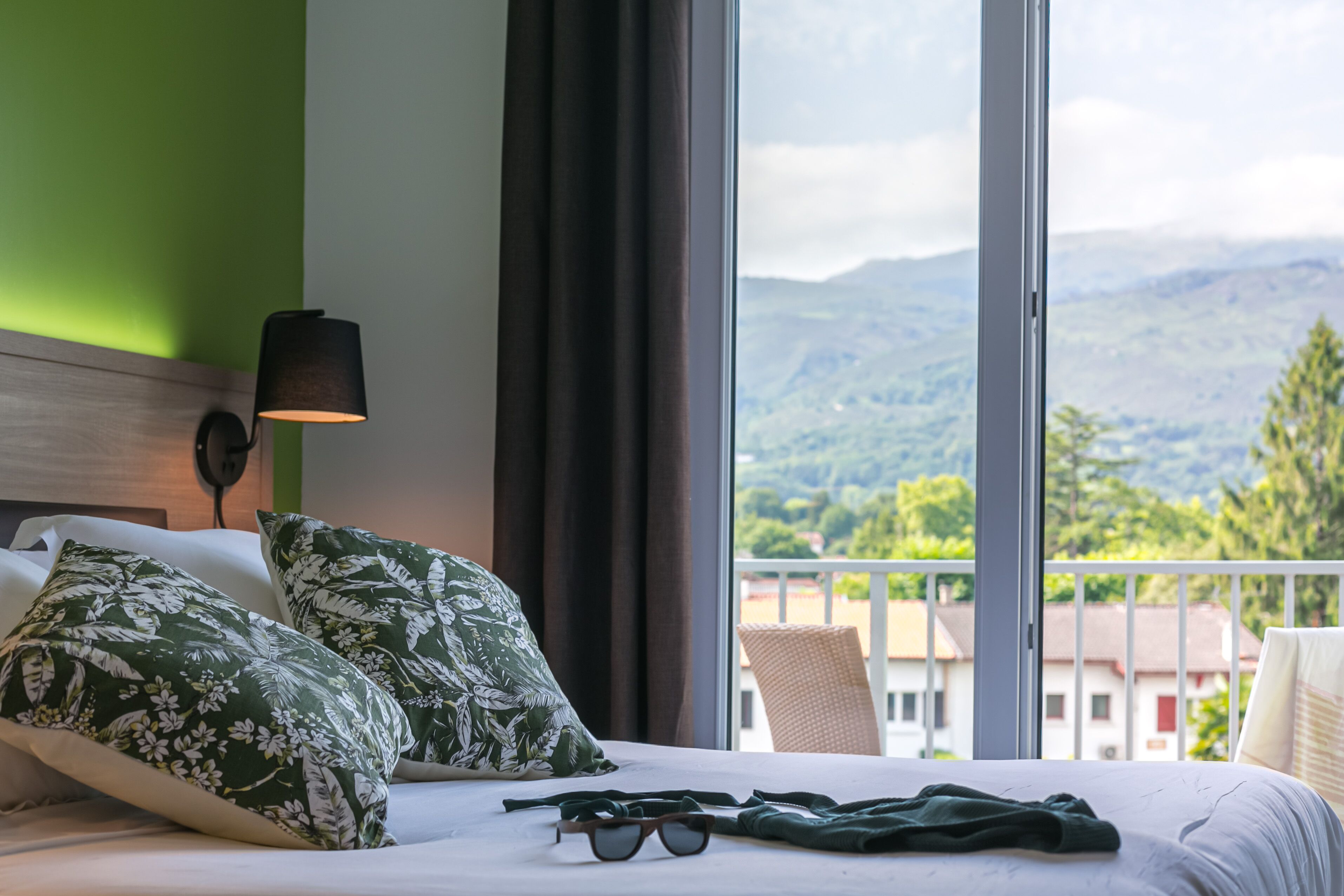 A hotel room with a bed, green patterned pillows, a pair of glasses, a lamp, and a view of mountains through large windows.