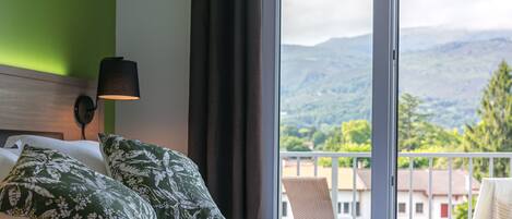 Habitación de hotel con una cama, almohadas de diseño verde, un par de gafas, una lámpara y vistas a montañas a través de amplios ventanales.