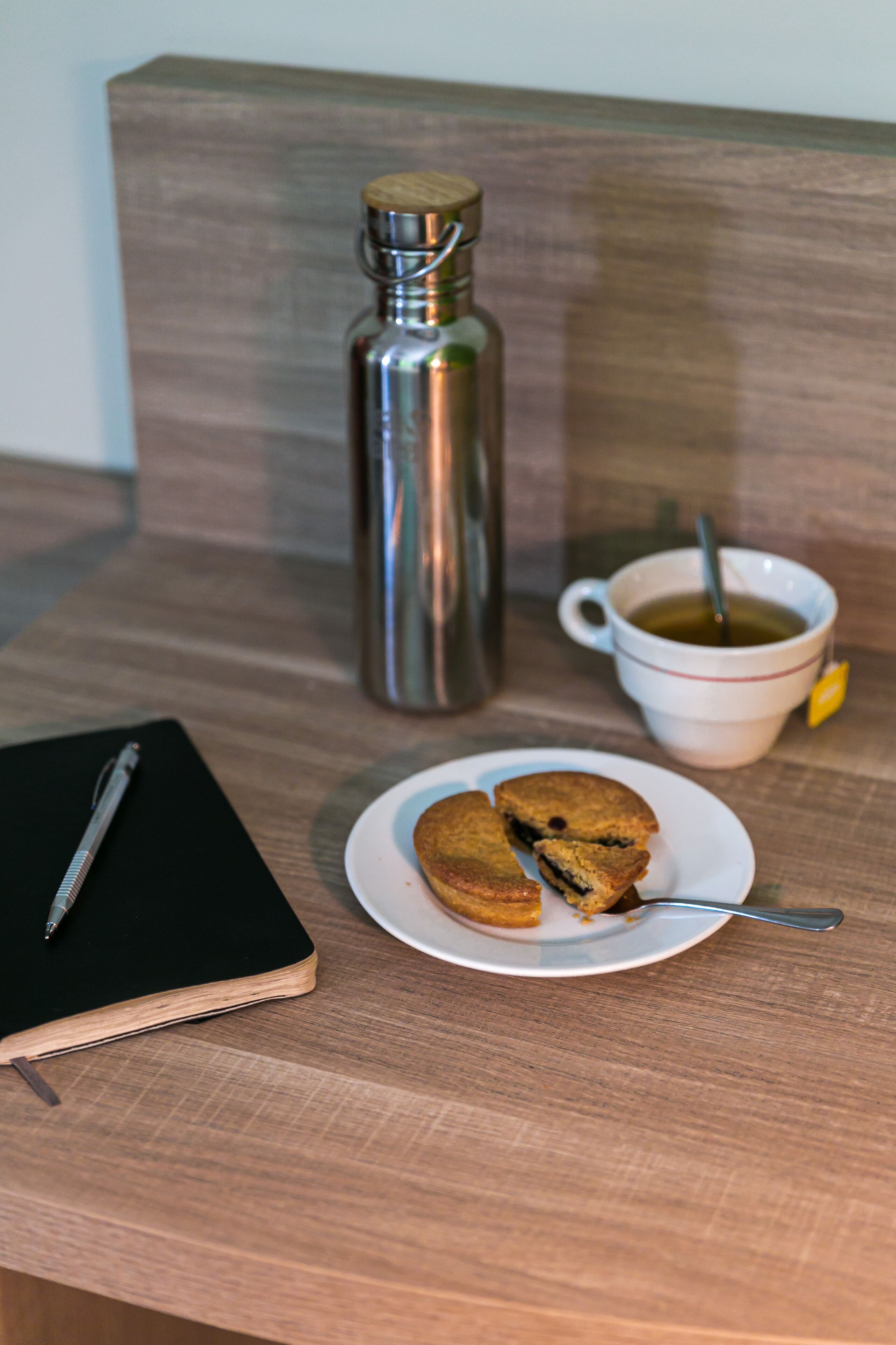 A wooden table with a notebook, a thermos, a mug with a spoon, and a plate with cookies.