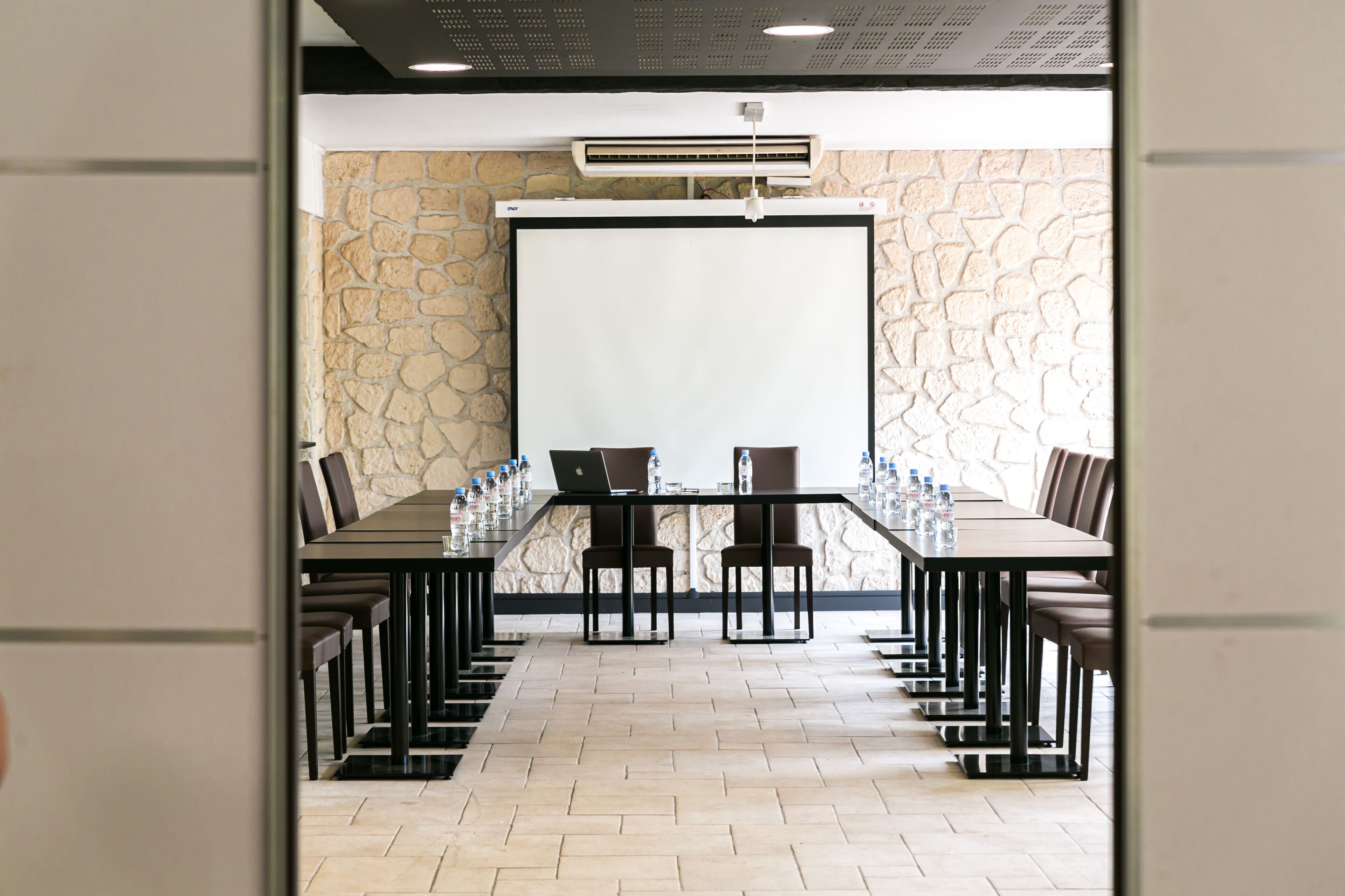 Een vergaderruimte met een lange tafel, stoelen, een projectiescherm en een airconditioning.
