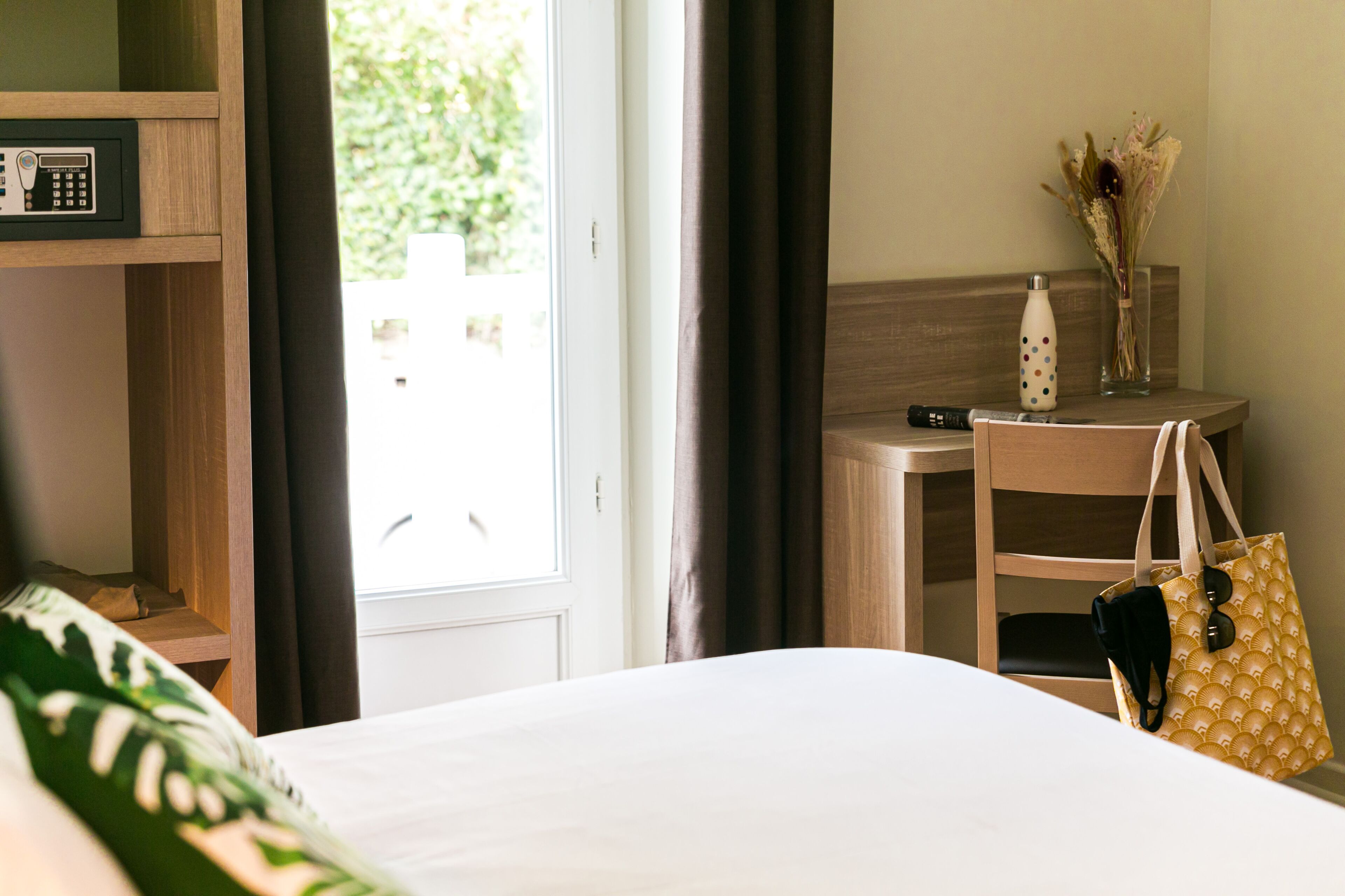 Een hotelkamer met een bed, een bureau met telefoon, een stoel, een klein tafeltje met een bloevas en een handtas op de stoel.