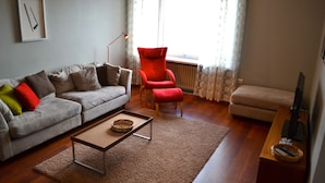 A living room with a grey sofa, red armchair, and beige ottoman. There is a coffee table with a basket, a floor lamp, and a TV stand with a remote. - Helsinki Apartment (Helsinki)
