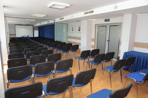 Un salón de conferencias con filas de sillas, una pantalla de proyector y una mesa azul.