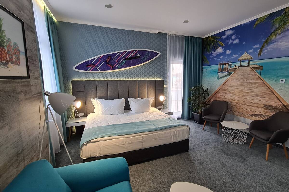 A hotel room with a bed, a desk, a chair, a small table, and a mural of a beach scene.