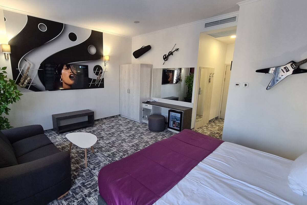 A modern hotel room with a large bed, a seating area with a sofa and a small table, a TV, a guitar, and a mirror on the wall.