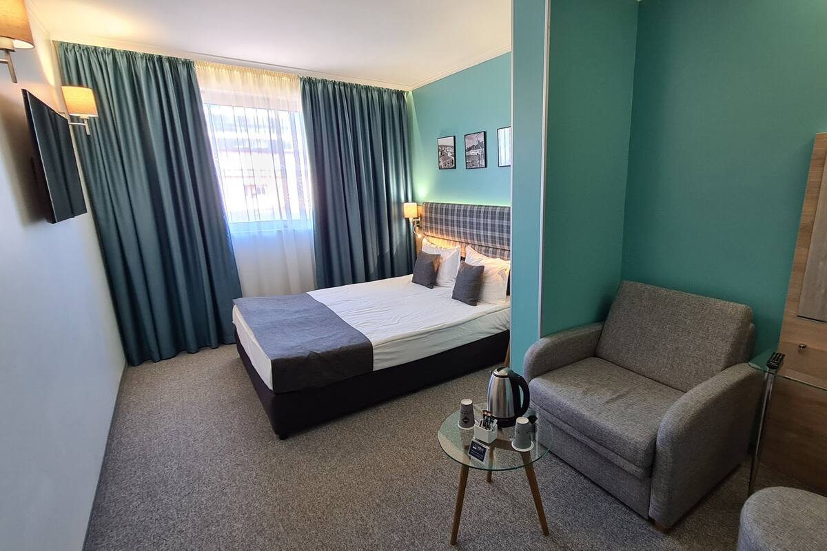 A hotel room with a bed, a sofa, a small table with a kettle and cups, a mirror, and a window with curtains.
