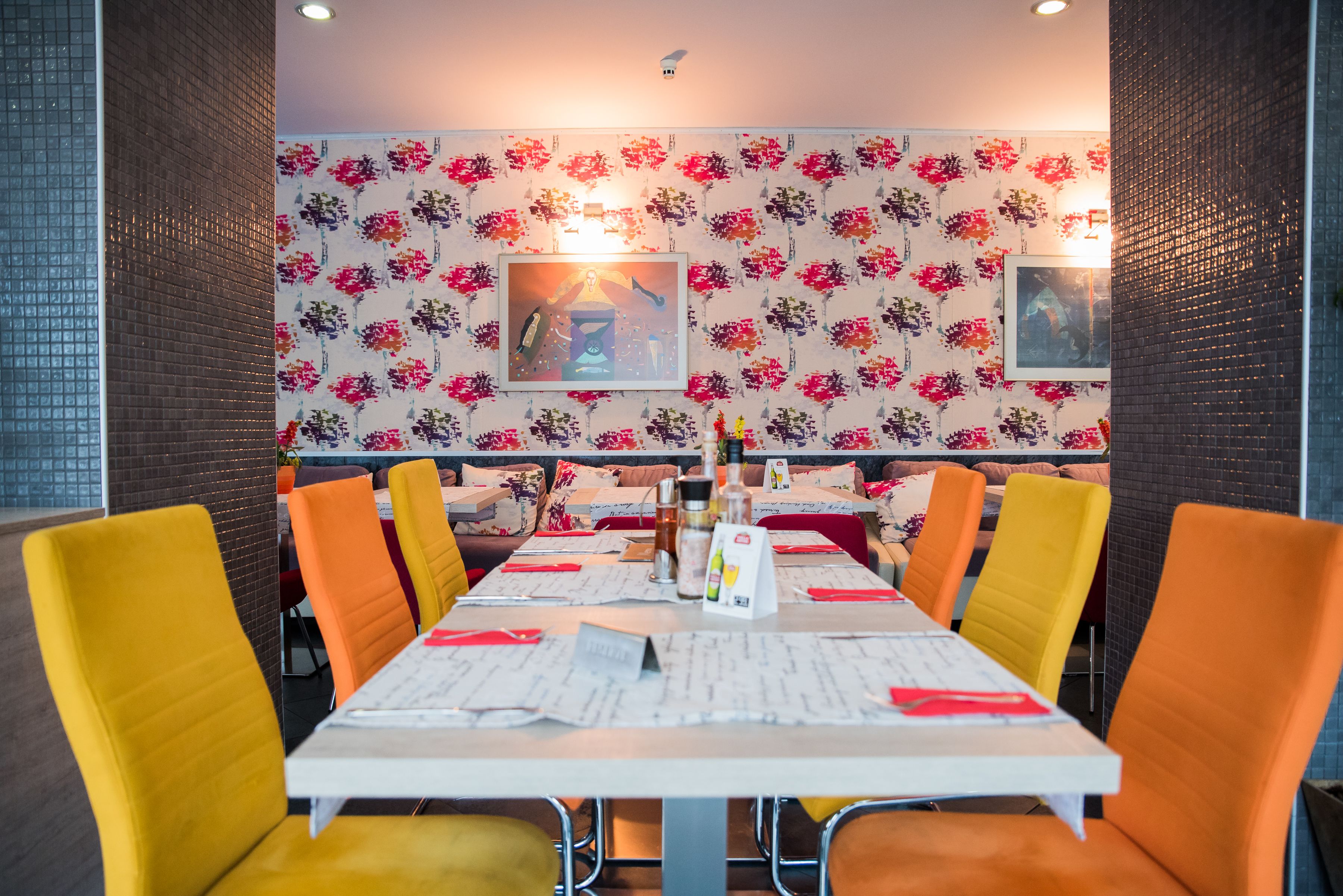Un restaurante con una larga mesa, sillas color naranja, papel tapiz floral y dos luces fijadas a la pared.