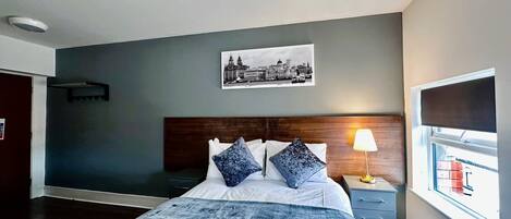 A bedroom with a bed, a nightstand, a lamp, a window, and a cityscape painting on the wall.