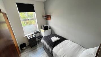 Un dormitorio pequeño y moderno con una cama individual, un escritorio con una computadora, una ventana con vistas a follaje y un mueble pequeño.