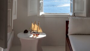 A room with a bed, a small table with a ship model, a window with a view of the sea, and a red rug on the floor.