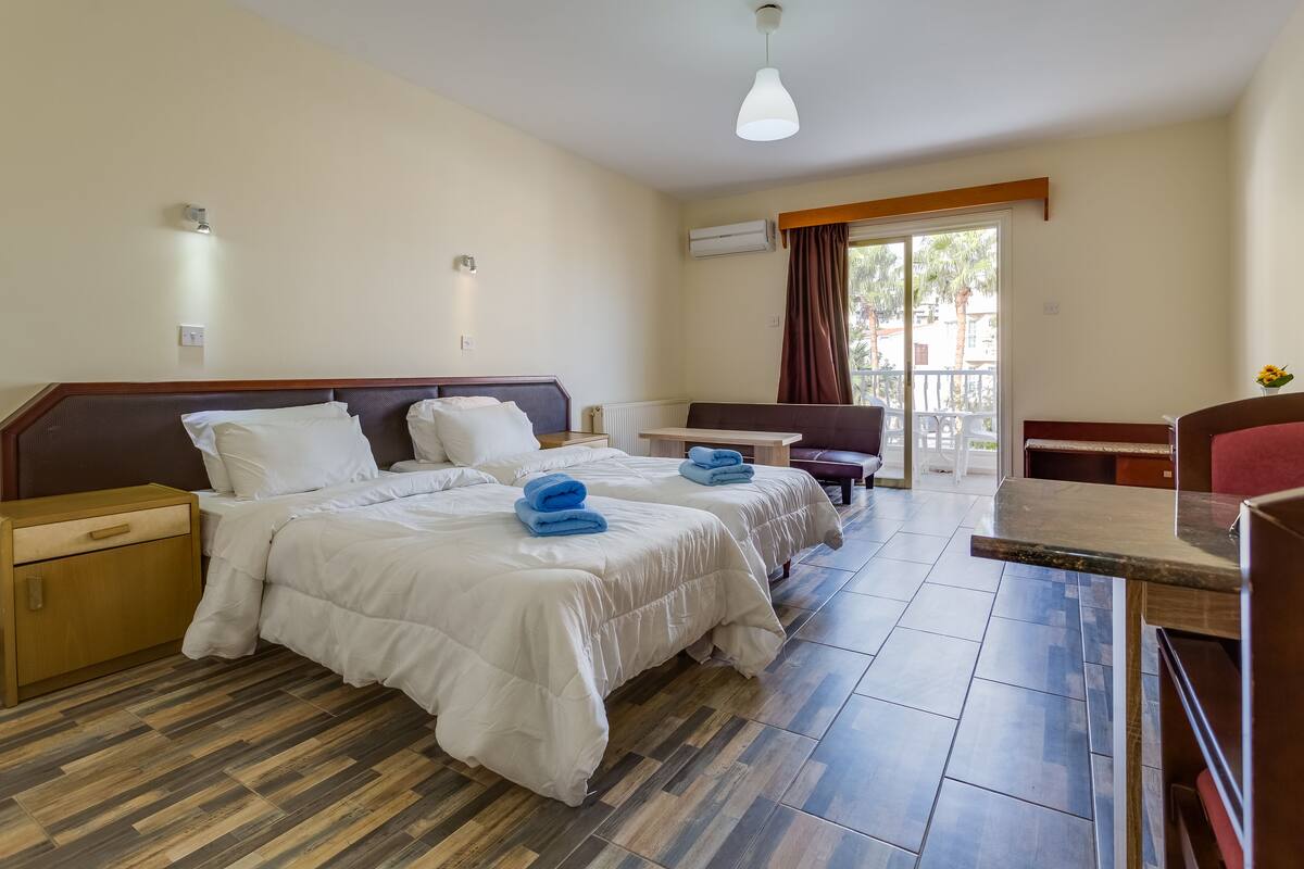 Quarto de hotel com duas camas, uma mesa de cabeceira de madeira, uma pequena mesa e uma varanda com vista.