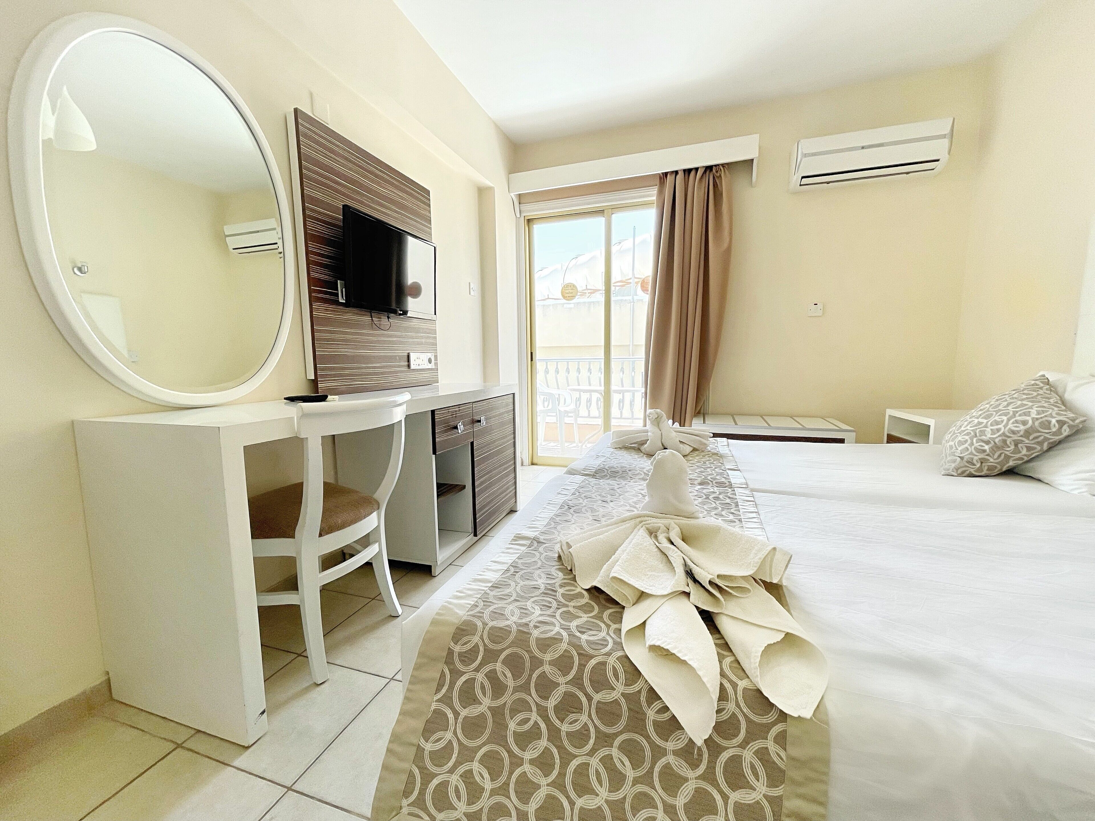 A hotel room with a bed, a desk with a mirror, a television, and an air conditioning unit.