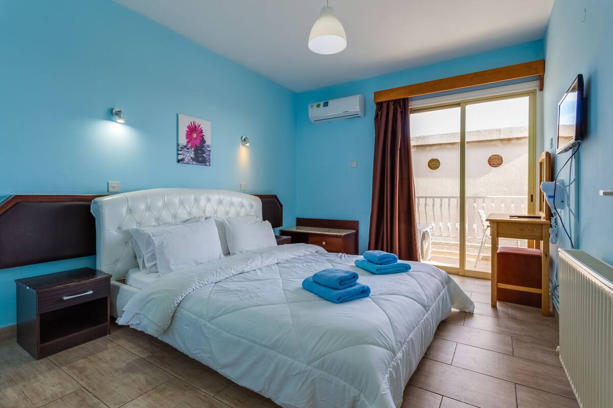 Quarto com cama, mesas de cabeceira, escrivaninha, cadeira e vista para uma varanda.