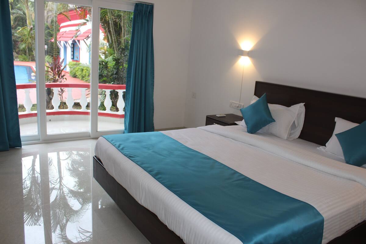 A hotel room with a large bed, a nightstand, and a view of a red-roofed building through a glass door.
