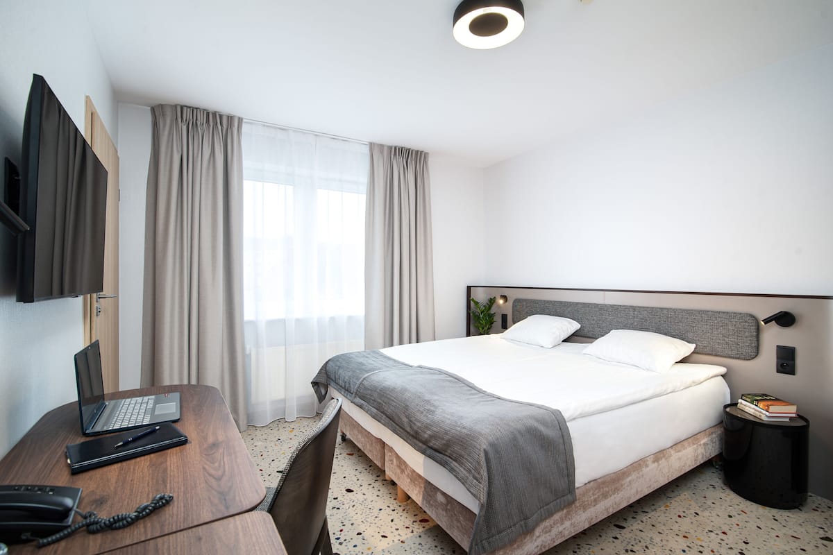 A hotel room with a bed, a desk with a laptop, a telephone, and a small side table with books.