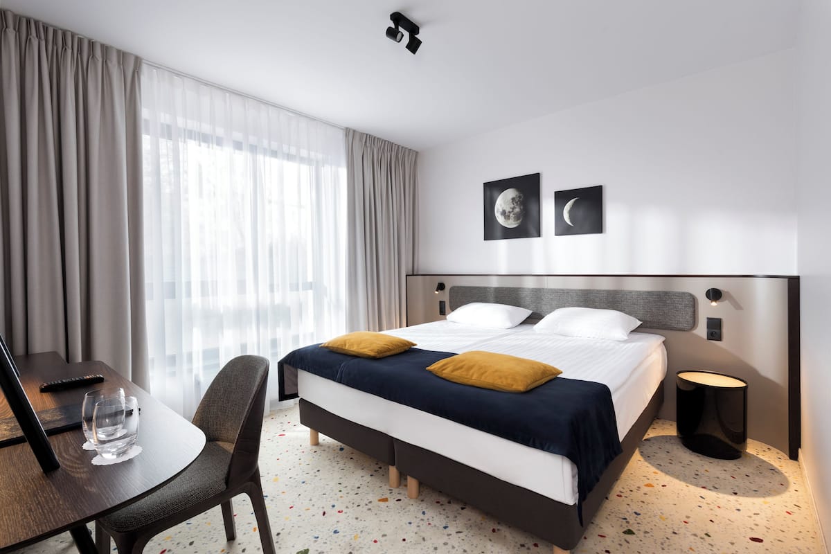 A hotel room with a bed, a desk, a chair, a bedside table, and a window with curtains.