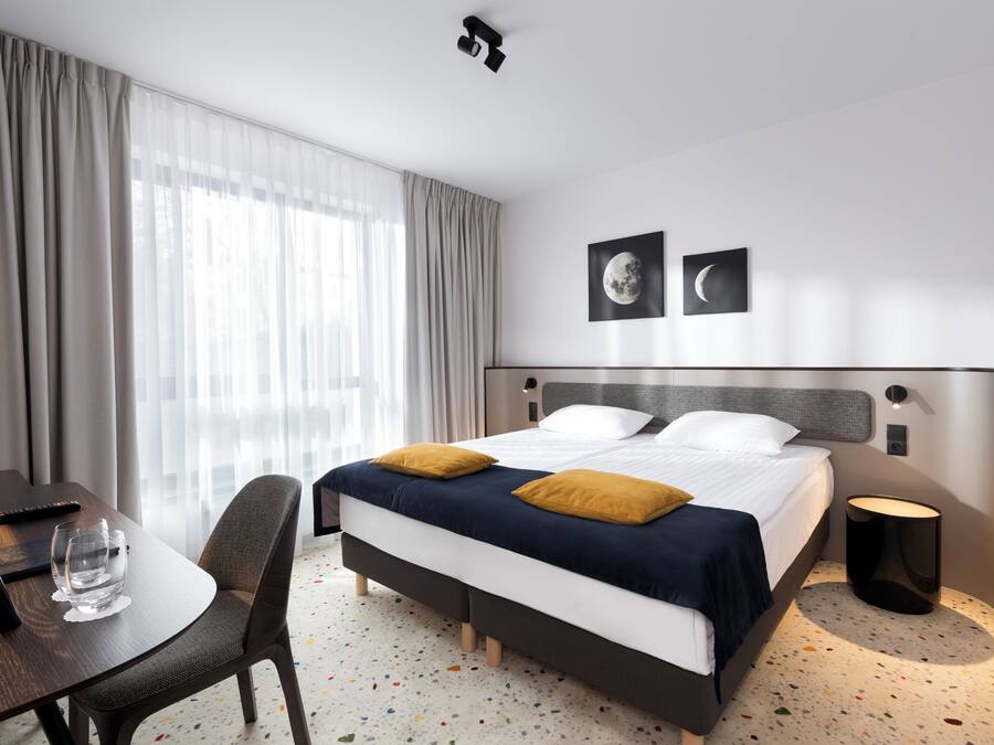 A hotel room with a bed, a desk, a chair, a bedside table, and a window with curtains.