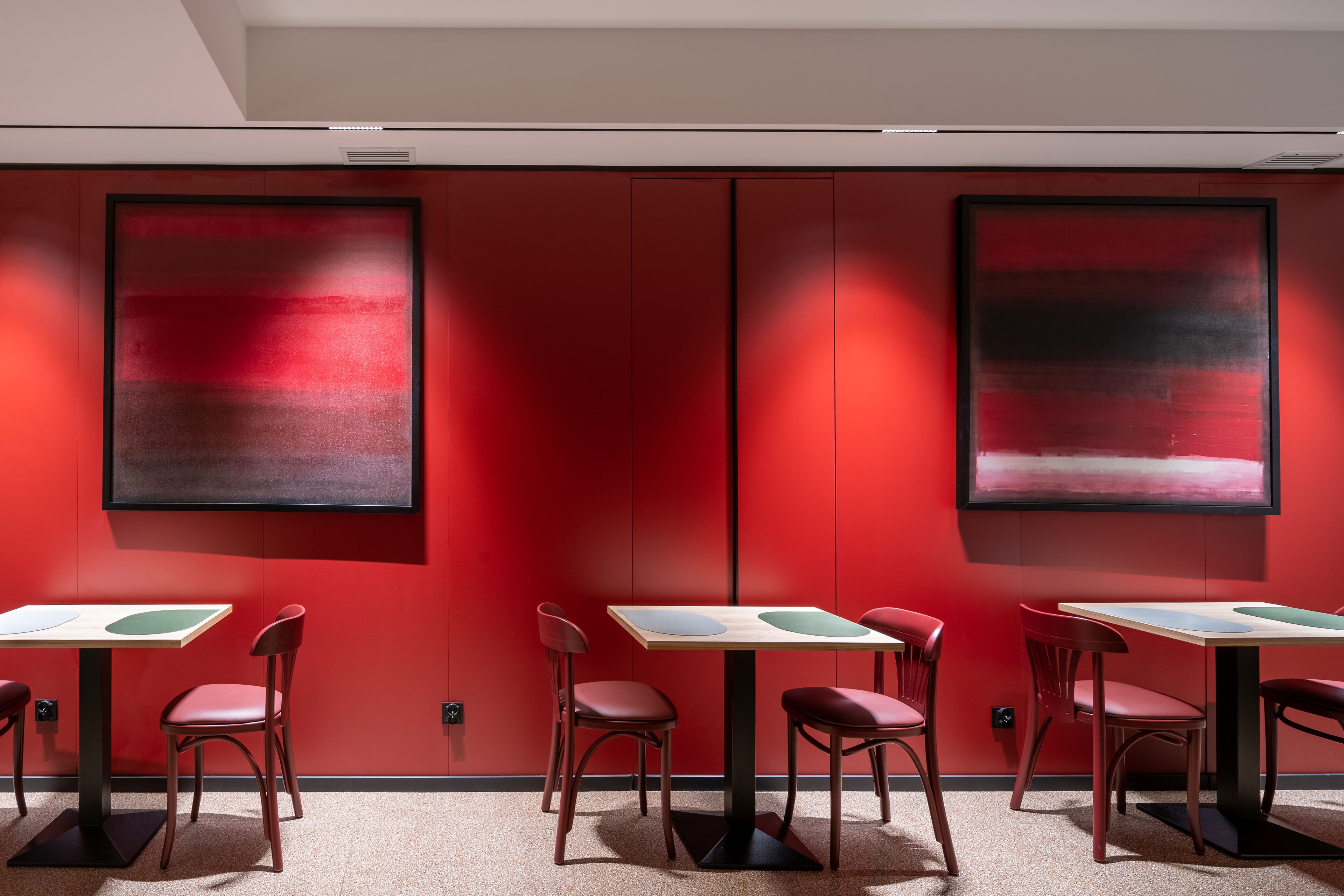 Una habitación moderna con paredes rojas, dos grandes pinturas abstractas y seis mesas con sillas.