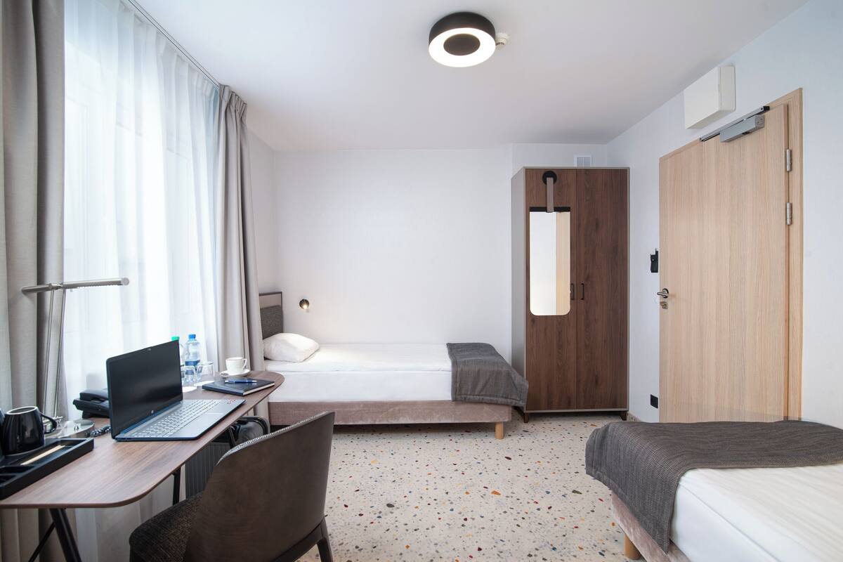 A hotel room with a bed, a desk with a laptop, a chair, a bedside table, a lamp, and a wardrobe.