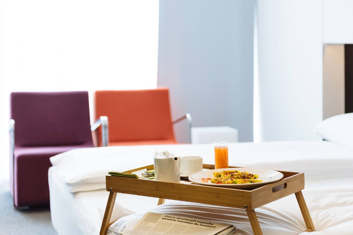 A hotel room with a bed, a wooden tray with breakfast items, a newspaper, and a red chair in the background.