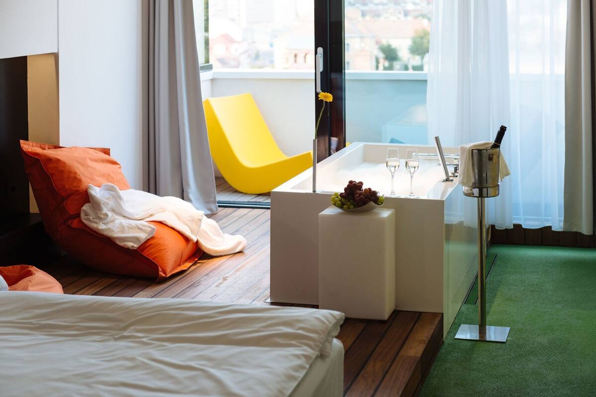 A modern hotel room with a bed, a yellow chair, a small white side table with a bowl of fruit, a bottle of champagne, and a green carpet.