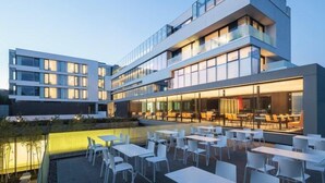 Een modern gebouw met meerdere verdiepingen, een terras met witte tafels en stoelen en een zwembad.