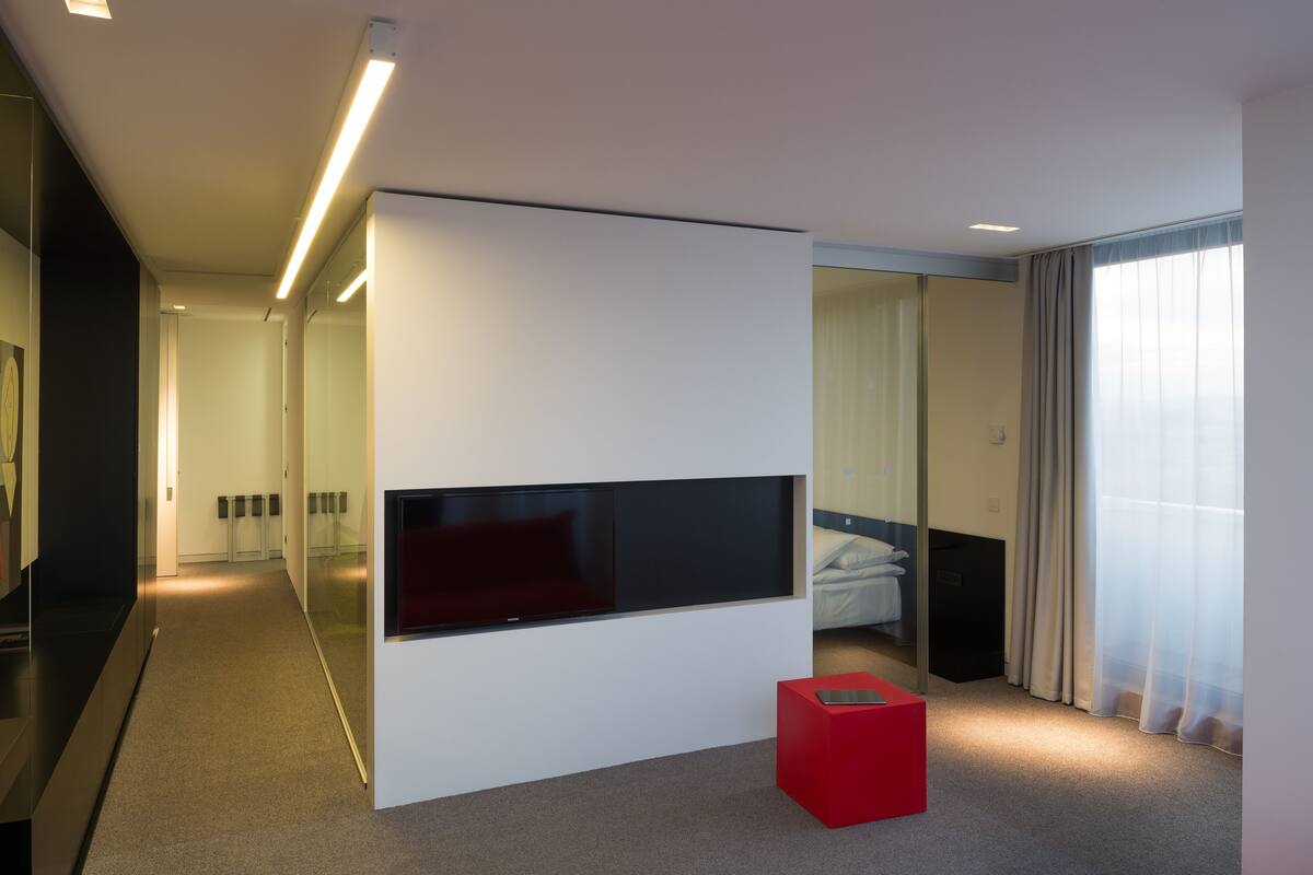 A modern hotel room with a large flat-screen TV mounted on a white wall, a red accent piece, and a large window with sheer curtains.
