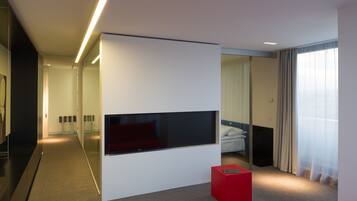 A modern hotel room with a large flat-screen TV mounted on a white wall, a red accent piece, and a large window with sheer curtains.