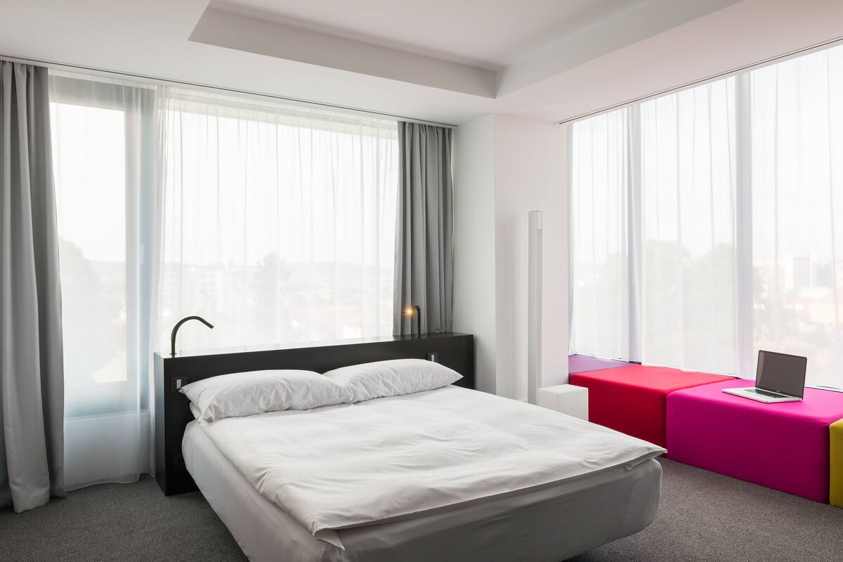 A modern hotel room with a large bed, a desk with a laptop, and a window with sheer curtains.