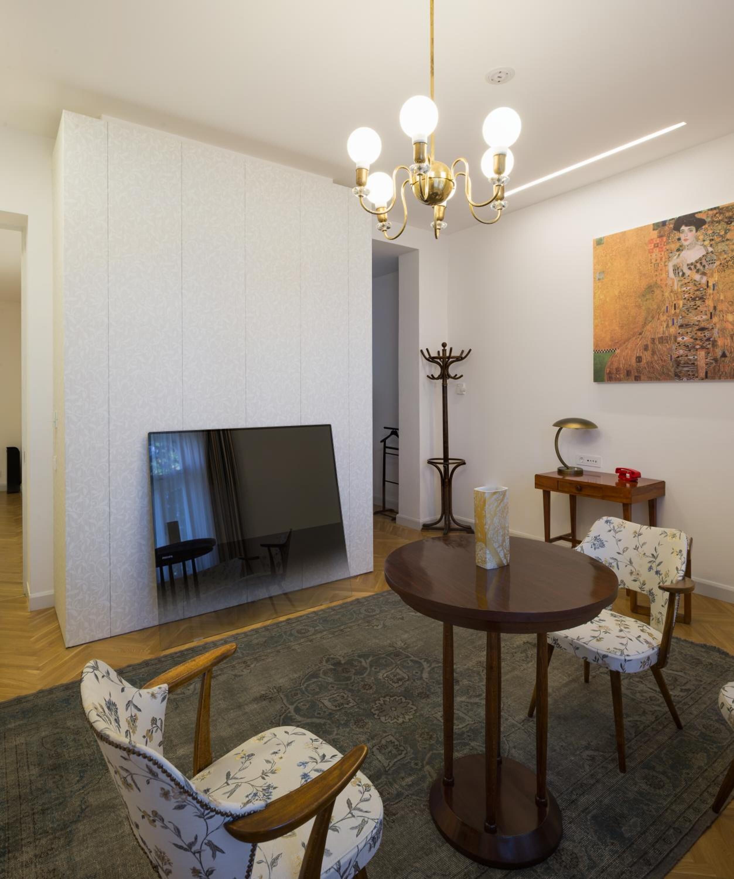 Un comedor con una mesa redonda, sillas con estampado floral, un candelabro, un perchero y un cuadro en la pared.