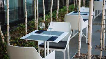 A dining area with white tables and chairs, arranged outdoors with birch trees and a glass wall in the background.
