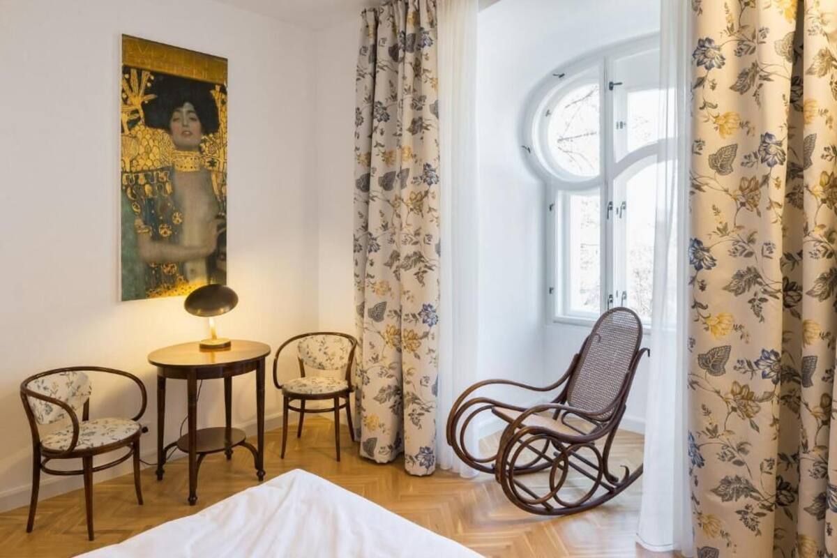 Een kamer met een bed, twee stoelen, een tafel met een lamp, een schilderij aan de muur en een raam met gordijnen.