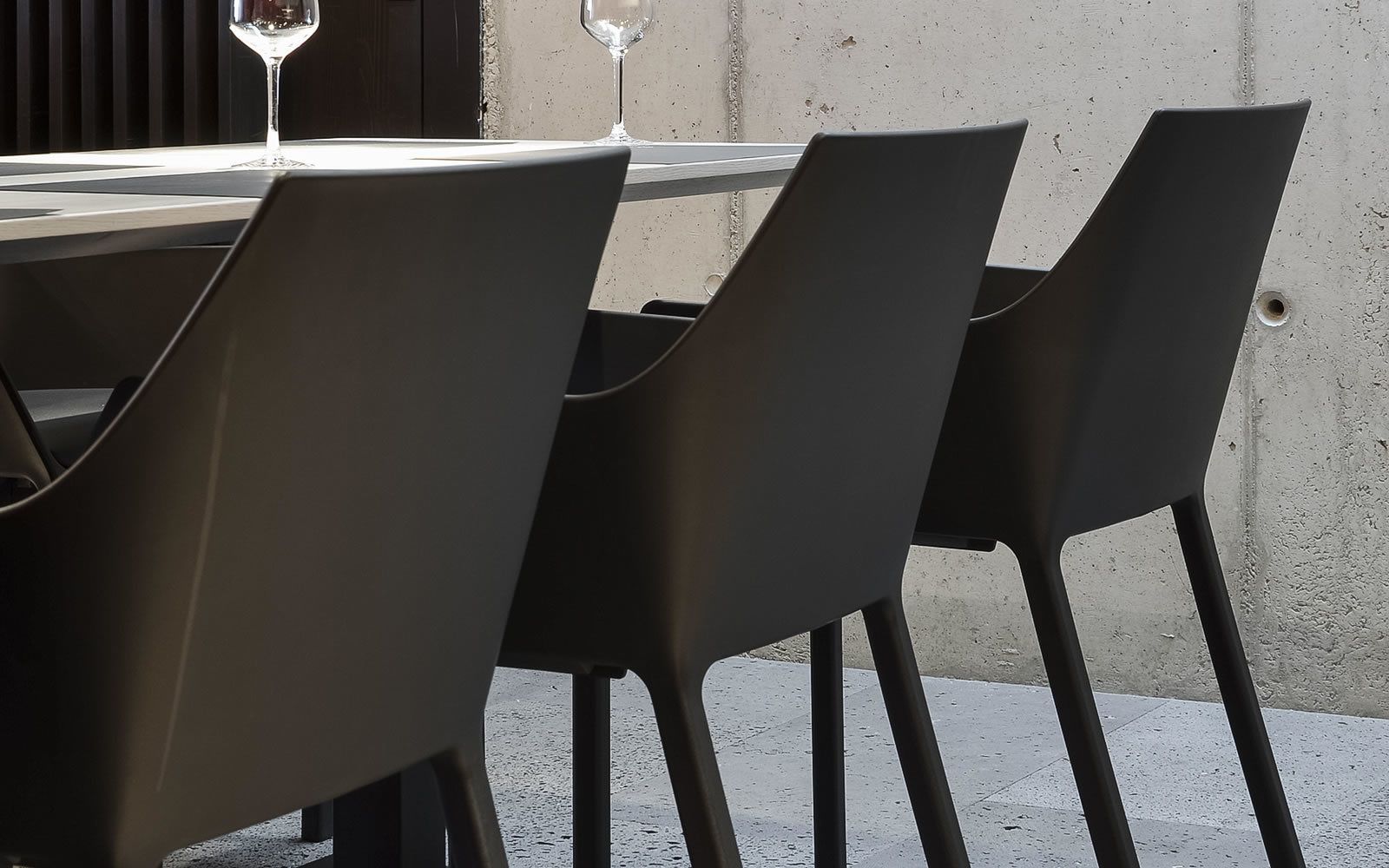 A table with four chairs, two wine glasses, and a concrete wall in the background.