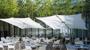 An outdoor dining area with white tables and chairs, large white umbrellas, and glass walls reflecting trees. - Villa Csonka by Hotel Privo (Targu Mures)
