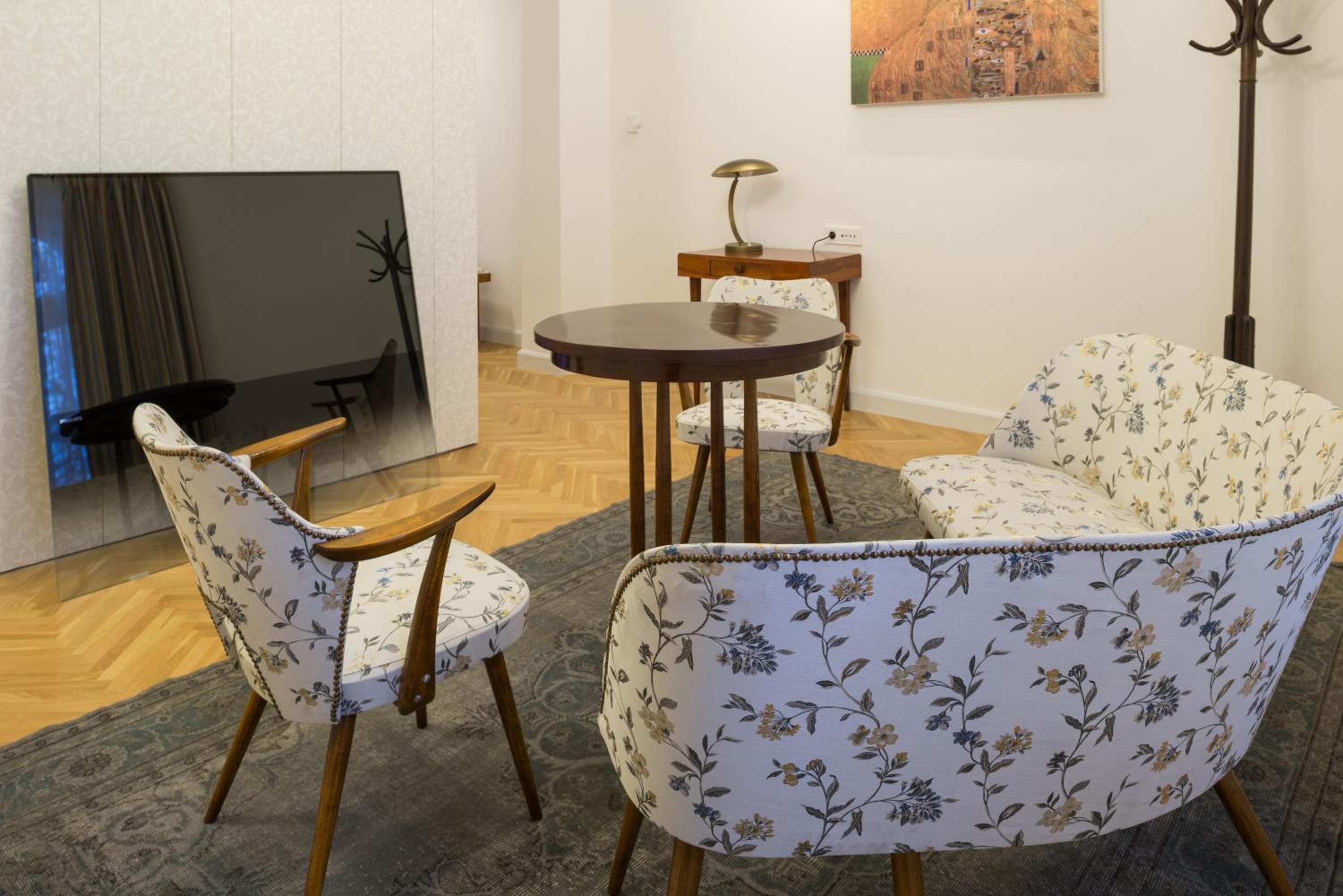 A living room with a round wooden table, two floral-patterned chairs, a floor lamp, and a flat-screen TV mounted on the wall.