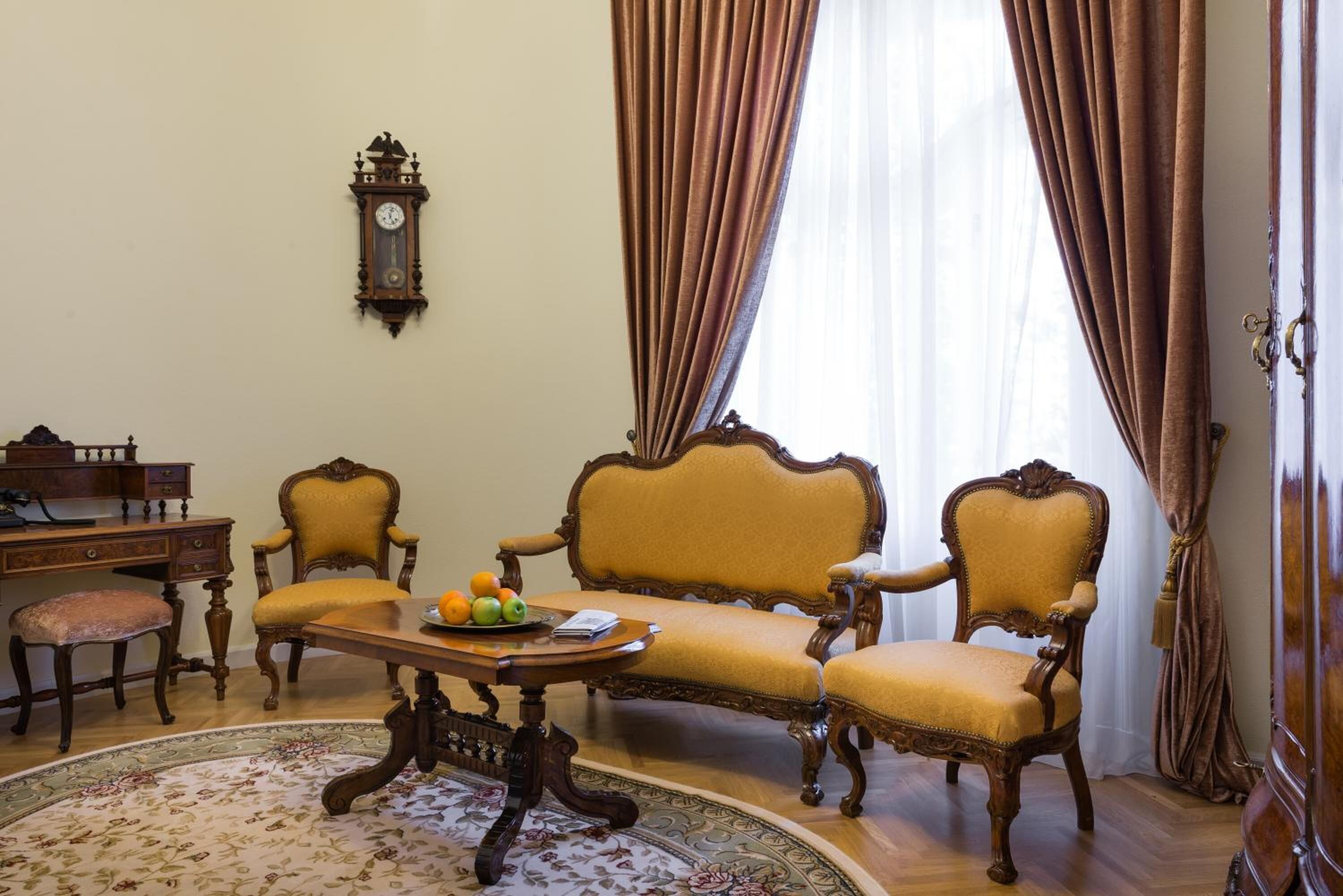 A room with a sofa, two armchairs, a wooden desk, a clock, and a table with fruit.