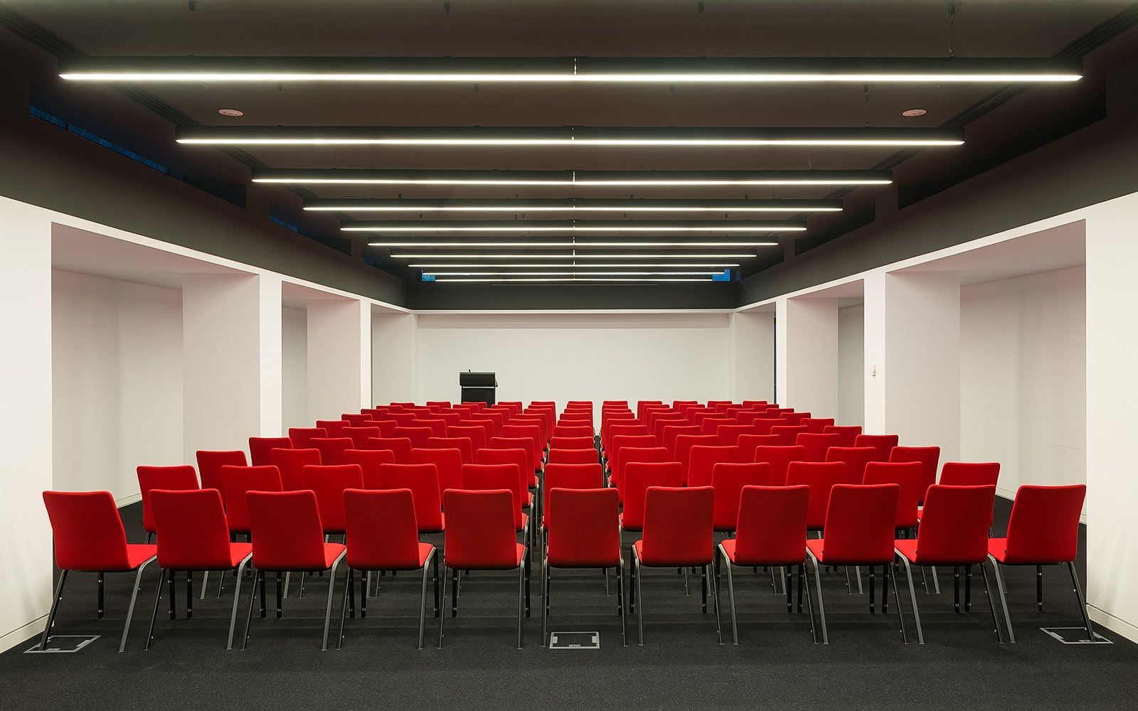 A conference room with rows of red chairs, a podium at the front, and modern lighting fixtures on the ceiling.