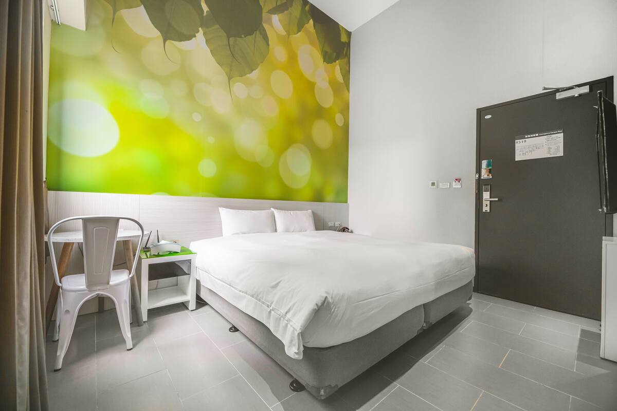 A modern hotel room with a large bed, a desk with a chair, and a door with a sign.