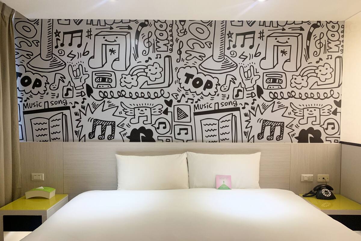A hotel room with a large bed, white pillows, and a wall decorated with black and white musical and book-themed drawings. There are two bedside tables, one with a green object and the other with a black object.