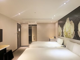 Habitación de hotel con dos camas, un escritorio, una silla, una mesita con cafetera y un cuadro floral en la pared.