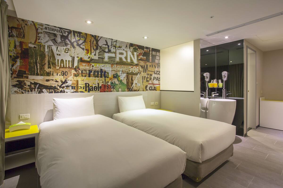 A hotel room with two beds, a large abstract mural on the wall, a mirror, and a bathroom with a sink and hairdryer.