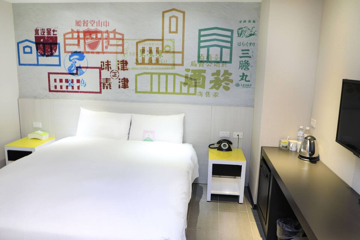 A hotel room with a bed, bedside tables, a TV, and a wall with colorful illustrations.