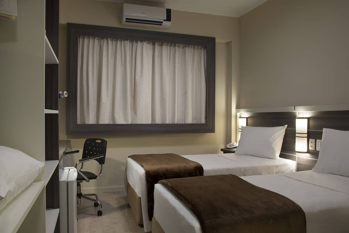 Habitación de hotel con dos camas, un escritorio con silla, aire acondicionado y una ventana con cortinas.