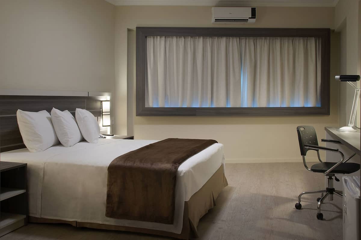 Una habitación de hotel con cama, mesita de noche, lámpara, escritorio, silla y aire acondicionado.