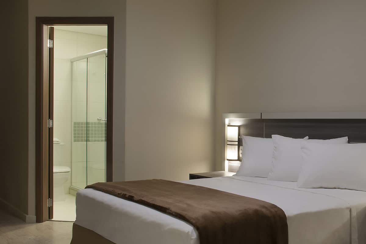 Una habitación de hotel con una cama grande, una mesita de noche y una zona de ducha con paredes de vidrio.