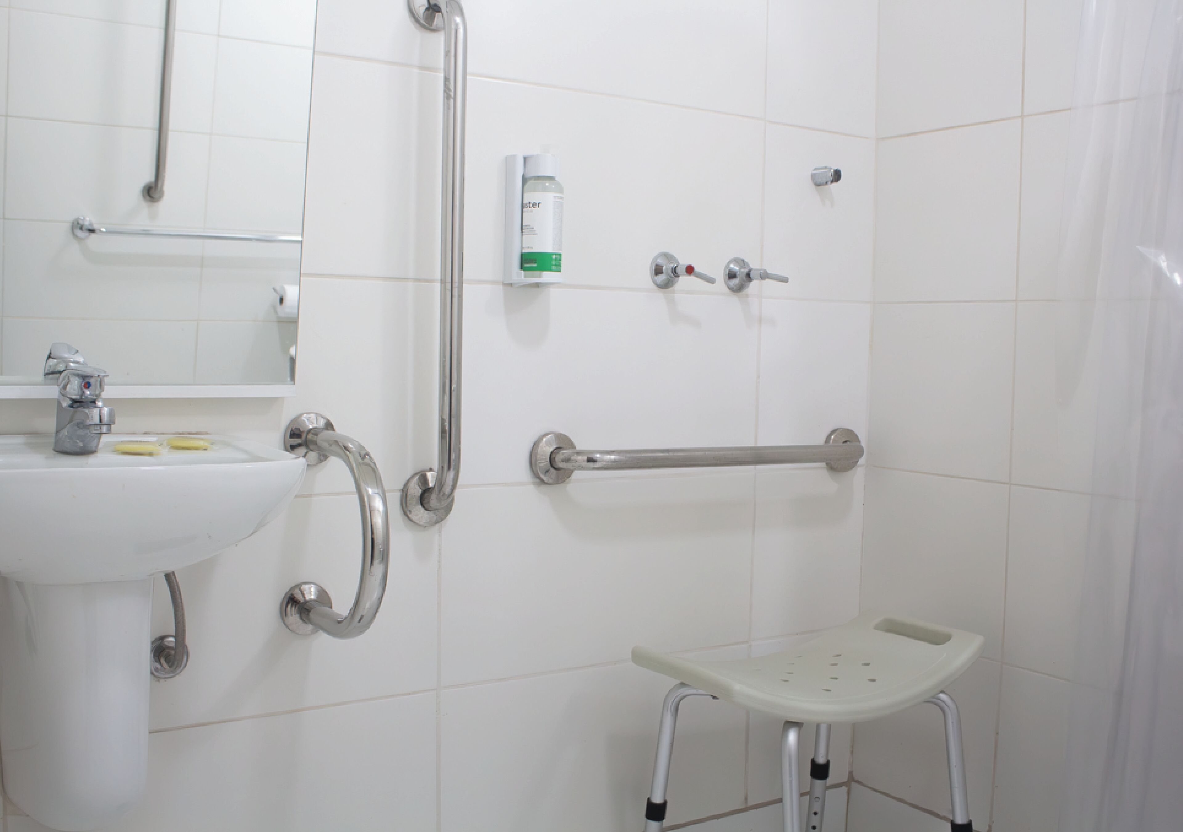 Un baño con lavamanos blanco, barras de apoyo, un espejo y un taburete pequeño.