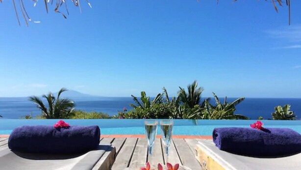 A wooden deck with two lounge chairs, each with a dark towel and a flower. Two glasses of champagne are on the deck, and a tropical view of the ocean is visible in the background. - Tet Rouge Resort St. Lucia (Choiseul)