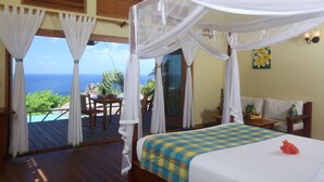 A bedroom with a bed, a balcony with a view of the ocean, and a dining area with a table and chairs. - Tet Rouge Resort St. Lucia (Choiseul)
