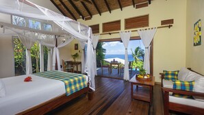 A room with a bed, a sofa, and a table with a fruit basket. There are windows with curtains and a view of the ocean. - Tet Rouge Resort St. Lucia (Choiseul)